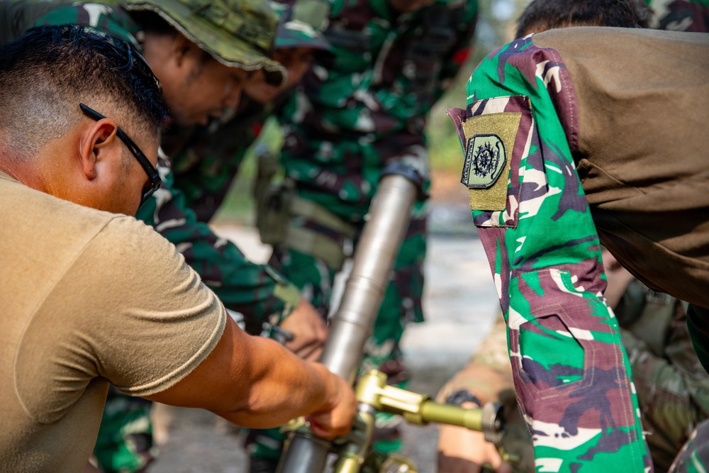 A group of military personnel are gathered around and handling what appears to be a mortar system. Most individuals are wearing green camouflage uniforms consistent with Indonesian Armed Forces (TNI) pattern, while one person on the left wears a tan/khaki t-shirt. The scene appears to be a training or demonstration exercise involving assembly or inspection of the weapon system.