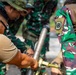 A group of military personnel are gathered around and handling what appears to be a mortar system. Most individuals are wearing green camouflage uniforms consistent with Indonesian Armed Forces (TNI) pattern, while one person on the left wears a tan/khaki t-shirt. The scene appears to be a training or demonstration exercise involving assembly or inspection of the weapon system.