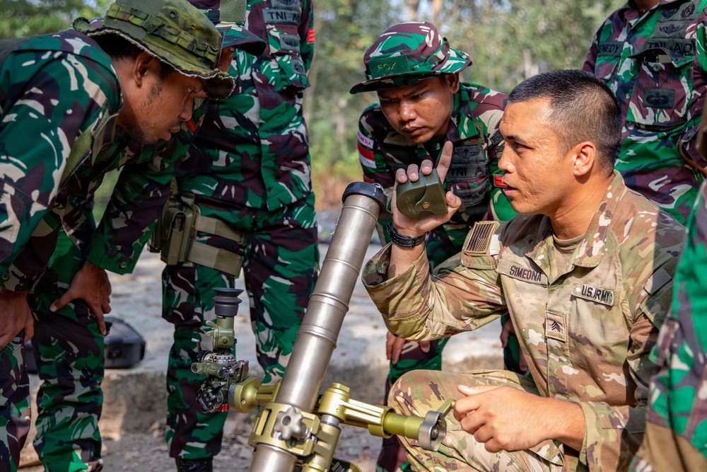 A U.S. Army soldier identified as 'SIMEONA' with the rank of Sergeant is conducting a training session with Indonesian military personnel (TNI - Tentara Nasional Indonesia). The soldier is demonstrating the operation of what appears to be an M224 60mm mortar system to a group of Indonesian soldiers in their distinctive green camouflage uniforms. This appears to be a joint military training exercise or bilateral cooperation event.