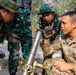A U.S. Army soldier identified as 'SIMEONA' with the rank of Sergeant is conducting a training session with Indonesian military personnel (TNI - Tentara Nasional Indonesia). The soldier is demonstrating the operation of what appears to be an M224 60mm mortar system to a group of Indonesian soldiers in their distinctive green camouflage uniforms. This appears to be a joint military training exercise or bilateral cooperation event.
