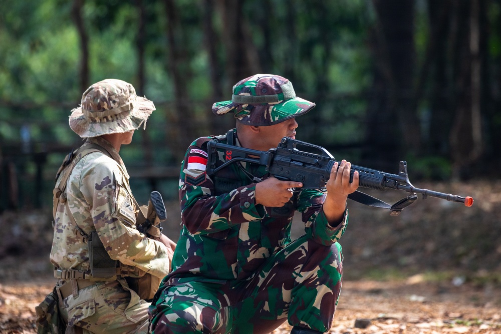 Two military personnel are engaged in what appears to be a joint training exercise in a forested outdoor environment. One soldier wearing Indonesian Army (TNI-AD) camouflage uniform with an 'INDONESIA' patch and number '32' is in a kneeling position aiming a rifle. A second soldier in a different camouflage pattern (MultiCam/OCP) with a tan boonie hat is positioned nearby, observing.