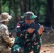 Two military personnel are engaged in what appears to be a joint training exercise in a forested outdoor environment. One soldier wearing Indonesian Army (TNI-AD) camouflage uniform with an 'INDONESIA' patch and number '32' is in a kneeling position aiming a rifle. A second soldier in a different camouflage pattern (MultiCam/OCP) with a tan boonie hat is positioned nearby, observing.