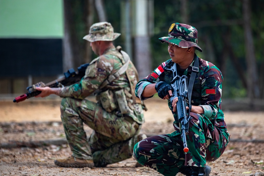 Two soldiers in different military uniforms are in kneeling tactical positions, each holding rifles. The soldier in the foreground wears green woodland camouflage with what appears to be an Indonesian flag patch, while the soldier in the background wears a MultiCam/OCP-style uniform. The setting appears to be an outdoor training area with dirt ground and trees in the background.
