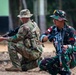 Two soldiers in different military uniforms are in kneeling tactical positions, each holding rifles. The soldier in the foreground wears green woodland camouflage with what appears to be an Indonesian flag patch, while the soldier in the background wears a MultiCam/OCP-style uniform. The setting appears to be an outdoor training area with dirt ground and trees in the background.