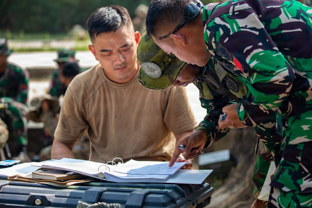 Two military personnel are reviewing documents in a binder placed on a hard-shell equipment case outdoors. One soldier wearing a tan t-shirt and green camouflage cap is seated, while another in green camouflage uniform with glasses leans over and points to a specific section of the paperwork. Additional military personnel are visible in the blurred background.