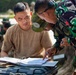 Two military personnel are reviewing documents in a binder placed on a hard-shell equipment case outdoors. One soldier wearing a tan t-shirt and green camouflage cap is seated, while another in green camouflage uniform with glasses leans over and points to a specific section of the paperwork. Additional military personnel are visible in the blurred background.