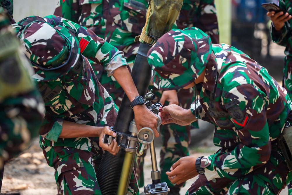 The image shows multiple soldiers in green-brown-white camouflage uniforms working together to assemble or adjust a mortar weapon system. The personnel appear to be conducting a training or demonstration exercise, with at least three soldiers actively handling the equipment. The camouflage pattern and uniform style are consistent with the Indonesian National Armed Forces (TNI).