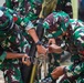 The image shows multiple soldiers in green-brown-white camouflage uniforms working together to assemble or adjust a mortar weapon system. The personnel appear to be conducting a training or demonstration exercise, with at least three soldiers actively handling the equipment. The camouflage pattern and uniform style are consistent with the Indonesian National Armed Forces (TNI).