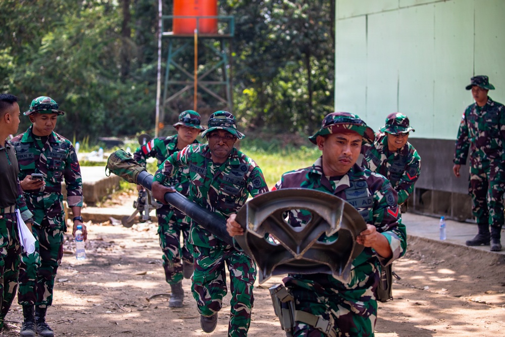 A group of military personnel wearing Indonesian Army (TNI-AD) camouflage uniforms and bush hats are engaged in what appears to be a training exercise or drill. Two soldiers in the foreground are running while carrying equipment, including what appears to be a tube/pipe and a metal frame component. The setting is an outdoor military training area with trees and a building in the background.