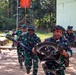 A group of military personnel wearing Indonesian Army (TNI-AD) camouflage uniforms and bush hats are engaged in what appears to be a training exercise or drill. Two soldiers in the foreground are running while carrying equipment, including what appears to be a tube/pipe and a metal frame component. The setting is an outdoor military training area with trees and a building in the background.