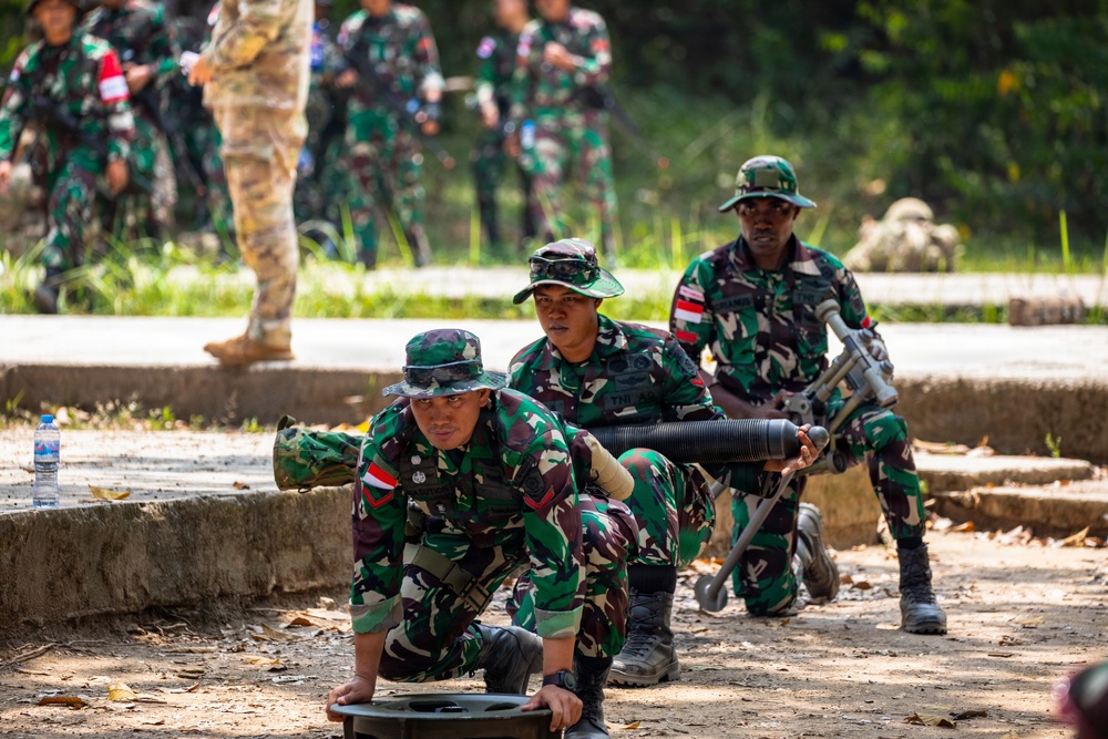 The image shows military personnel in green camouflage uniforms conducting what appears to be a training exercise or demonstration. Three soldiers in the foreground are handling what appears to be a mortar system, with one soldier positioning the base plate and another managing the tube. Additional soldiers and at least one person in a different camouflage pattern (possibly an advisor or observer) are visible in the background.