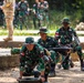 The image shows military personnel in green camouflage uniforms conducting what appears to be a training exercise or demonstration. Three soldiers in the foreground are handling what appears to be a mortar system, with one soldier positioning the base plate and another managing the tube. Additional soldiers and at least one person in a different camouflage pattern (possibly an advisor or observer) are visible in the background.