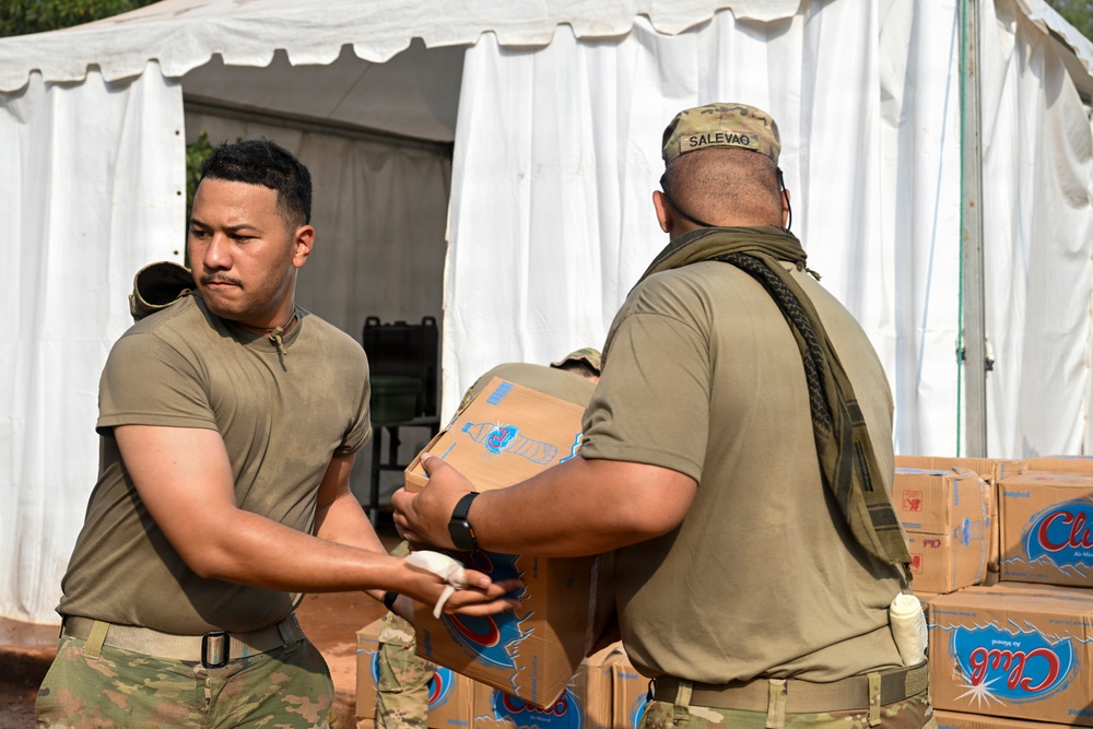 Two military personnel in olive/multicam uniforms are handling cardboard boxes, likely during a supply or humanitarian aid distribution operation near a white tent structure. The soldier facing away has a name tape reading 'SALEVAO' on his uniform. Stacked boxes of what appears to be bottled water (Club brand) are visible in the background.