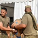Two military personnel in olive/multicam uniforms are handling cardboard boxes, likely during a supply or humanitarian aid distribution operation near a white tent structure. The soldier facing away has a name tape reading 'SALEVAO' on his uniform. Stacked boxes of what appears to be bottled water (Club brand) are visible in the background.