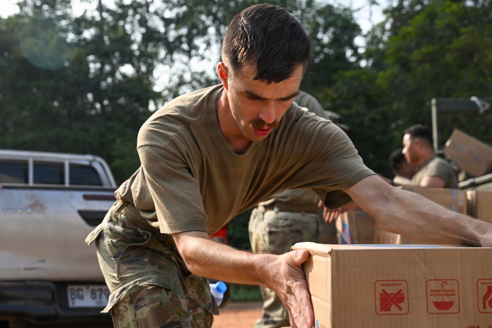 A military service member in olive drab t-shirt and camouflage pants is handling a cardboard box, likely during a logistics or humanitarian aid operation. Additional personnel in similar uniforms are visible in the background also handling boxes. The setting appears to be an outdoor area with red dirt ground, trees, and a pickup truck visible.