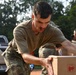 A military service member in olive drab t-shirt and camouflage pants is handling a cardboard box, likely during a logistics or humanitarian aid operation. Additional personnel in similar uniforms are visible in the background also handling boxes. The setting appears to be an outdoor area with red dirt ground, trees, and a pickup truck visible.