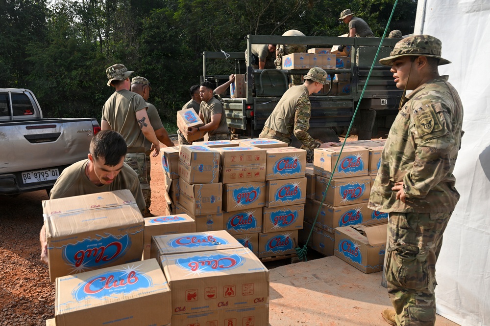 Military personnel in camouflage uniforms are unloading and stacking cardboard boxes labeled 'Club' from a military truck onto a dirt surface near a white tent structure. The operation appears to be a humanitarian aid or supply distribution mission conducted in a tropical or subtropical outdoor environment. Multiple soldiers are actively engaged in the logistics effort, with some on the truck bed and others organizing boxes on the ground.