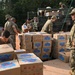 Military personnel in camouflage uniforms are unloading and stacking cardboard boxes labeled 'Club' from a military truck onto a dirt surface near a white tent structure. The operation appears to be a humanitarian aid or supply distribution mission conducted in a tropical or subtropical outdoor environment. Multiple soldiers are actively engaged in the logistics effort, with some on the truck bed and others organizing boxes on the ground.