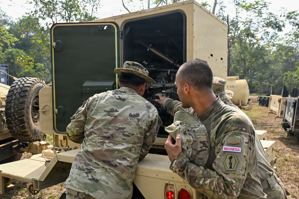 Two soldiers in multicam/OCP camouflage uniforms are examining the interior mechanical components of a military vehicle, likely during a joint training exercise. One soldier has a name tape reading 'BARAYUGA' and the other has a country tape reading 'INDONESIA' on his right sleeve, along with a unit patch. They appear to be conducting maintenance or an inspection of the vehicle's engine or equipment compartment in an outdoor wooded area.