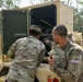 Two soldiers in multicam/OCP camouflage uniforms are examining the interior mechanical components of a military vehicle, likely during a joint training exercise. One soldier has a name tape reading 'BARAYUGA' and the other has a country tape reading 'INDONESIA' on his right sleeve, along with a unit patch. They appear to be conducting maintenance or an inspection of the vehicle's engine or equipment compartment in an outdoor wooded area.