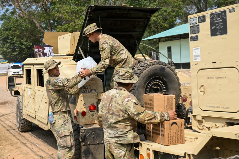Three soldiers in MultiCam/OCP camouflage uniforms are loading or unloading supplies from a tan military HMMWV (Humvee). The vehicle is marked with 'MSC 303 MEB' and the operation appears to take place at a military installation with a banner visible in the background containing text in what appears to be Indonesian ('Terus Melak... Samp...'). Cardboard boxes, likely MREs or similar military rations, and a clear plastic bag are being handled.