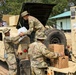 Three soldiers in MultiCam/OCP camouflage uniforms are loading or unloading supplies from a tan military HMMWV (Humvee). The vehicle is marked with 'MSC 303 MEB' and the operation appears to take place at a military installation with a banner visible in the background containing text in what appears to be Indonesian ('Terus Melak... Samp...'). Cardboard boxes, likely MREs or similar military rations, and a clear plastic bag are being handled.
