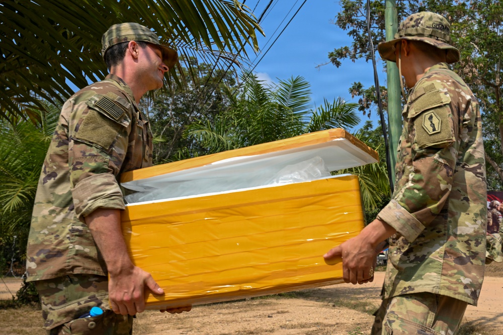 Two soldiers in MultiCam/OCP camouflage uniforms are carrying a large yellow cooler or insulated container together in an outdoor tropical setting. The soldier on the left displays a US flag patch on his right shoulder, while the soldier on the right has a name tape reading 'GUZMAN' and a unit patch visible. The setting appears to be a humanitarian aid or medical supply distribution operation in a tropical region.