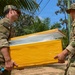 Two soldiers in MultiCam/OCP camouflage uniforms are carrying a large yellow cooler or insulated container together in an outdoor tropical setting. The soldier on the left displays a US flag patch on his right shoulder, while the soldier on the right has a name tape reading 'GUZMAN' and a unit patch visible. The setting appears to be a humanitarian aid or medical supply distribution operation in a tropical region.
