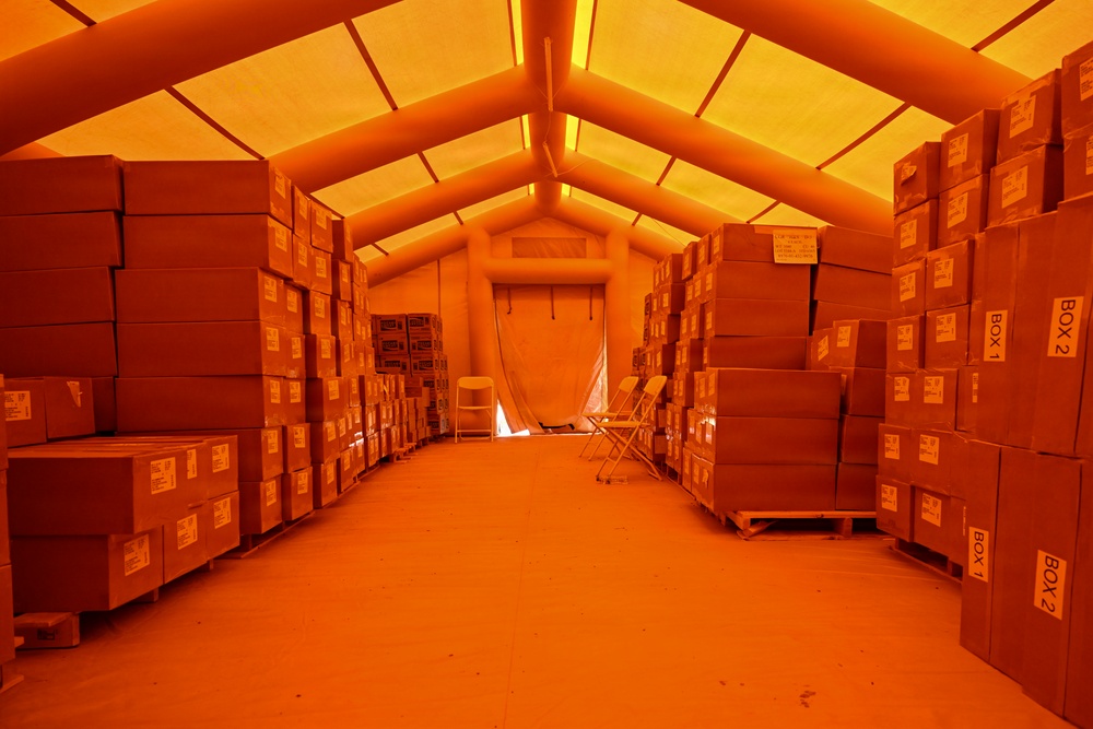 The image shows the interior of an inflatable or tensioned fabric structure used as a temporary warehouse or storage facility. Rows of cardboard boxes stacked on wooden pallets line both sides of a central aisle, extending toward a zippered fabric door at the far end. The entire scene is bathed in a deep orange/amber light, likely from the tent material filtering external light or internal orange lighting.