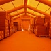 The image shows the interior of an inflatable or tensioned fabric structure used as a temporary warehouse or storage facility. Rows of cardboard boxes stacked on wooden pallets line both sides of a central aisle, extending toward a zippered fabric door at the far end. The entire scene is bathed in a deep orange/amber light, likely from the tent material filtering external light or internal orange lighting.