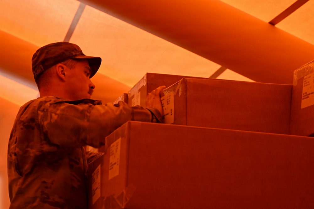 A person wearing military camouflage uniform and cap is handling or inspecting cardboard boxes in what appears to be a cargo hold or storage area. The entire scene is bathed in an intense orange-red light, likely from an aircraft or vehicle interior lighting. The individual appears to be stacking, organizing, or checking the boxes.