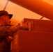 A person wearing military camouflage uniform and cap is handling or inspecting cardboard boxes in what appears to be a cargo hold or storage area. The entire scene is bathed in an intense orange-red light, likely from an aircraft or vehicle interior lighting. The individual appears to be stacking, organizing, or checking the boxes.
