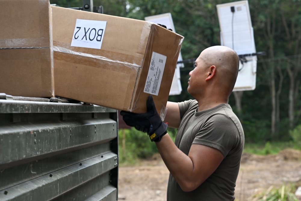 A bald man wearing an olive/military green t-shirt and black tactical gloves is loading or unloading a cardboard box labeled 'BOX 2' onto or from a military-style vehicle or equipment rack. The setting appears to be an outdoor area with trees in the background, suggesting a field or operational environment. The image captures a logistics or supply handling activity.