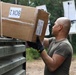 A bald man wearing an olive/military green t-shirt and black tactical gloves is loading or unloading a cardboard box labeled 'BOX 2' onto or from a military-style vehicle or equipment rack. The setting appears to be an outdoor area with trees in the background, suggesting a field or operational environment. The image captures a logistics or supply handling activity.