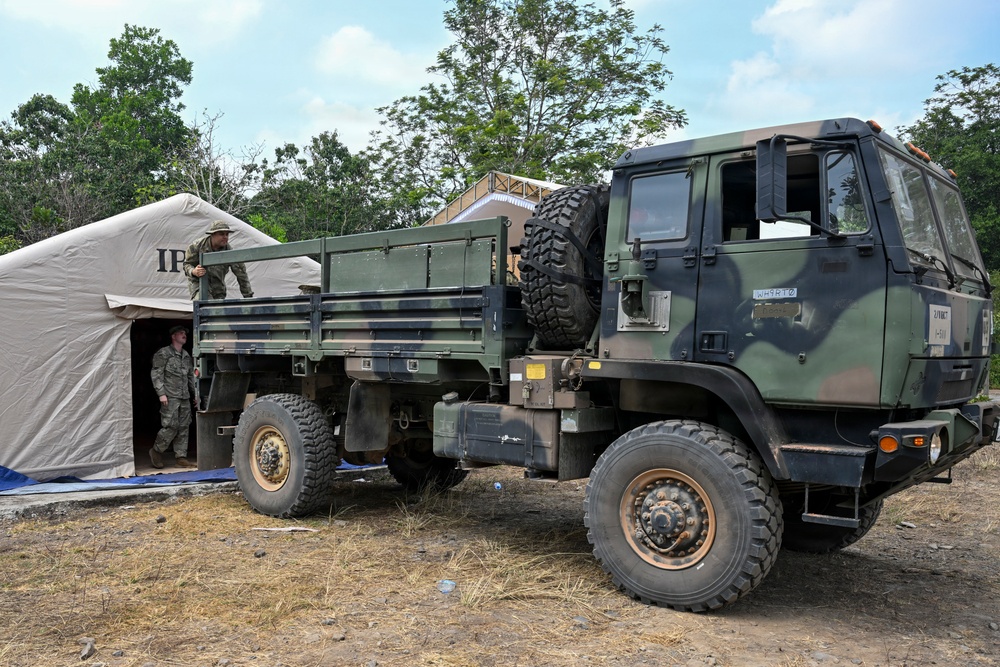 A military field operation showing a camouflage-painted tactical truck (appears to be an LMTV or similar light medium tactical vehicle) parked adjacent to a tan military tent. Two personnel in military uniforms are visible, one standing in the truck bed and one near the tent entrance. The setting appears to be a forward operating base or field camp in a tropical or subtropical environment.