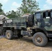 A military field operation showing a camouflage-painted tactical truck (appears to be an LMTV or similar light medium tactical vehicle) parked adjacent to a tan military tent. Two personnel in military uniforms are visible, one standing in the truck bed and one near the tent entrance. The setting appears to be a forward operating base or field camp in a tropical or subtropical environment.