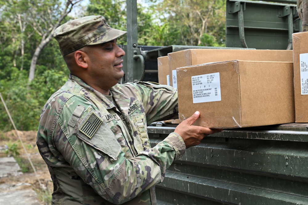 A U.S. Army soldier in OCP (Operational Camouflage Pattern) uniform is handling cardboard boxes on what appears to be a military vehicle or loading dock in a wooded outdoor environment. The boxes appear to be MRE (Meals Ready to Eat) ration cases based on visible labeling. The soldier appears to be loading or unloading supplies.