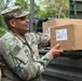 A U.S. Army soldier in OCP (Operational Camouflage Pattern) uniform is handling cardboard boxes on what appears to be a military vehicle or loading dock in a wooded outdoor environment. The boxes appear to be MRE (Meals Ready to Eat) ration cases based on visible labeling. The soldier appears to be loading or unloading supplies.