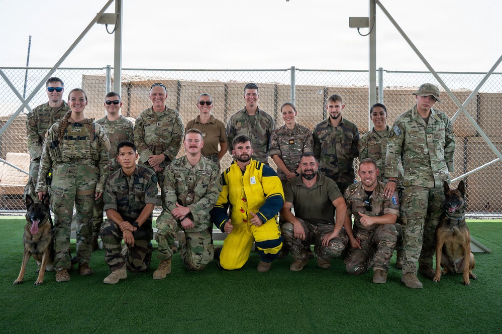 A group of approximately 17 military personnel in camouflage uniforms pose for a group photo on an artificial turf surface, likely at a forward operating base or military installation. Two military working dogs (Belgian Malinois) are present on either side of the group. One individual in the front center is wearing a distinctive yellow and blue bite/protection suit, commonly used in military working dog training exercises.