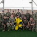 A group of approximately 17 military personnel in camouflage uniforms pose for a group photo on an artificial turf surface, likely at a forward operating base or military installation. Two military working dogs (Belgian Malinois) are present on either side of the group. One individual in the front center is wearing a distinctive yellow and blue bite/protection suit, commonly used in military working dog training exercises.