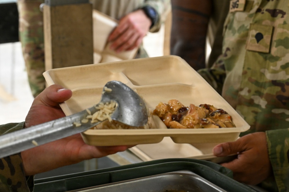Military personnel are being served a meal in a field or dining facility setting. A soldier is holding a compartmentalized biodegradable food tray while another uses a large serving spoon to portion out food. The tray contains what appears to be a glazed chicken dish with dried fruit and a noodle or rice side dish.