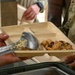 Military personnel are being served a meal in a field or dining facility setting. A soldier is holding a compartmentalized biodegradable food tray while another uses a large serving spoon to portion out food. The tray contains what appears to be a glazed chicken dish with dried fruit and a noodle or rice side dish.