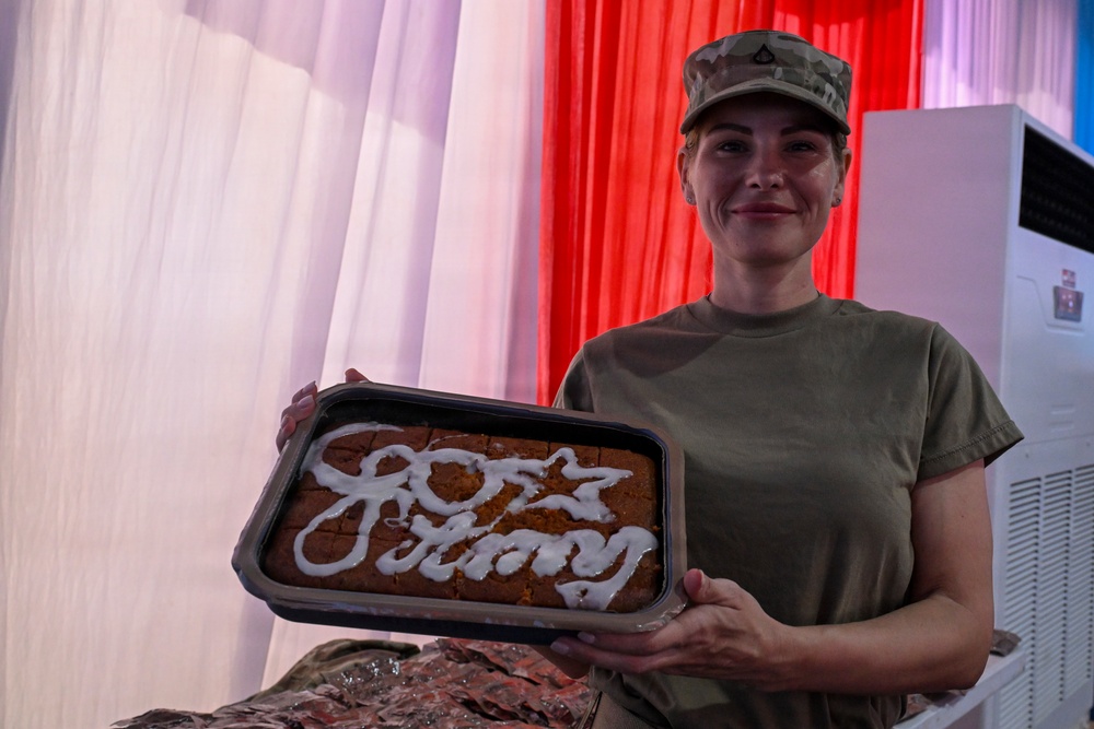 A person in military uniform (camouflage cap and olive green t-shirt) is smiling and holding a baking pan containing a decorated baked good. The baked item appears to have white icing writing on it, likely reading 'Army' along with a number or symbol. The background features red, white, and pink curtains suggesting a celebratory or ceremonial setting.