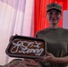 A person in military uniform (camouflage cap and olive green t-shirt) is smiling and holding a baking pan containing a decorated baked good. The baked item appears to have white icing writing on it, likely reading 'Army' along with a number or symbol. The background features red, white, and pink curtains suggesting a celebratory or ceremonial setting.