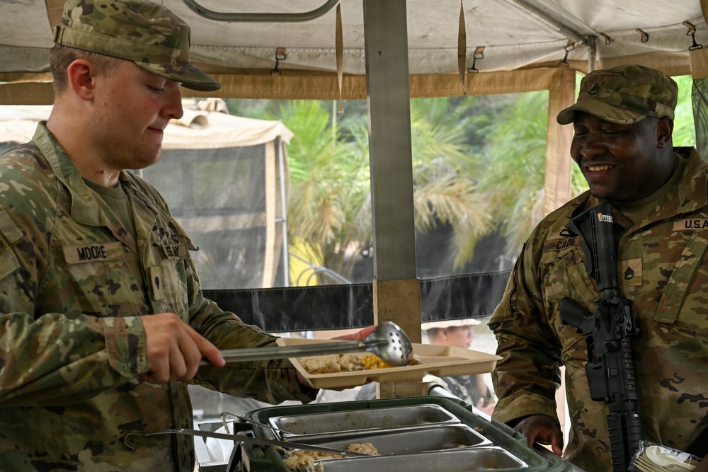 Two U.S. Army soldiers are at a field mess tent, with one soldier (name tape: MOORE) serving food to another soldier (name tape: CADE) who is smiling. The setting appears to be an outdoor military field kitchen under a canvas tent structure. The soldier being served is carrying a rifle and bears Staff Sergeant rank insignia.