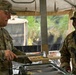 Two U.S. Army soldiers are at a field mess tent, with one soldier (name tape: MOORE) serving food to another soldier (name tape: CADE) who is smiling. The setting appears to be an outdoor military field kitchen under a canvas tent structure. The soldier being served is carrying a rifle and bears Staff Sergeant rank insignia.