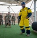 A military working dog (MWD) training exercise is underway at an outdoor facility. A decoy/agitator wearing a yellow and blue bite protection suit and blue helmet is positioned in front of a handler holding a Belgian Malinois on leash. A group of military personnel in MultiCam/OCP uniforms observe the demonstration from the background.