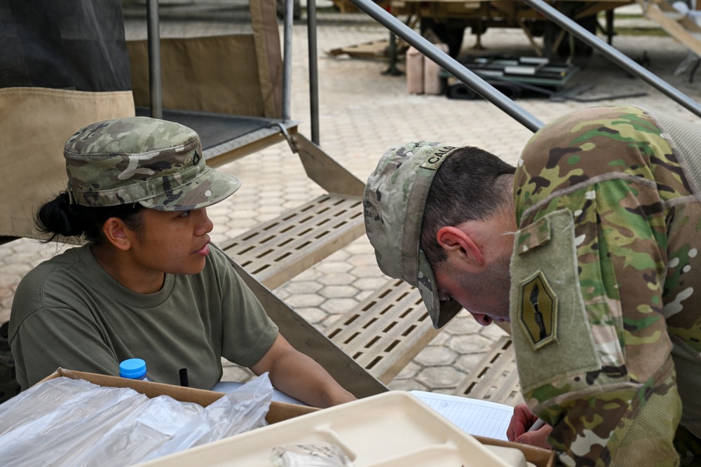 Two U.S. Army soldiers in Operational Camouflage Pattern (OCP) uniforms are engaged in an administrative or logistical task at an outdoor field station. One soldier is seated behind a table with documents and packaged items, while the other is leaning over to review or sign paperwork. The setting appears to be a forward operating base or field exercise environment.