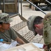 Two U.S. Army soldiers in Operational Camouflage Pattern (OCP) uniforms are engaged in an administrative or logistical task at an outdoor field station. One soldier is seated behind a table with documents and packaged items, while the other is leaning over to review or sign paperwork. The setting appears to be a forward operating base or field exercise environment.