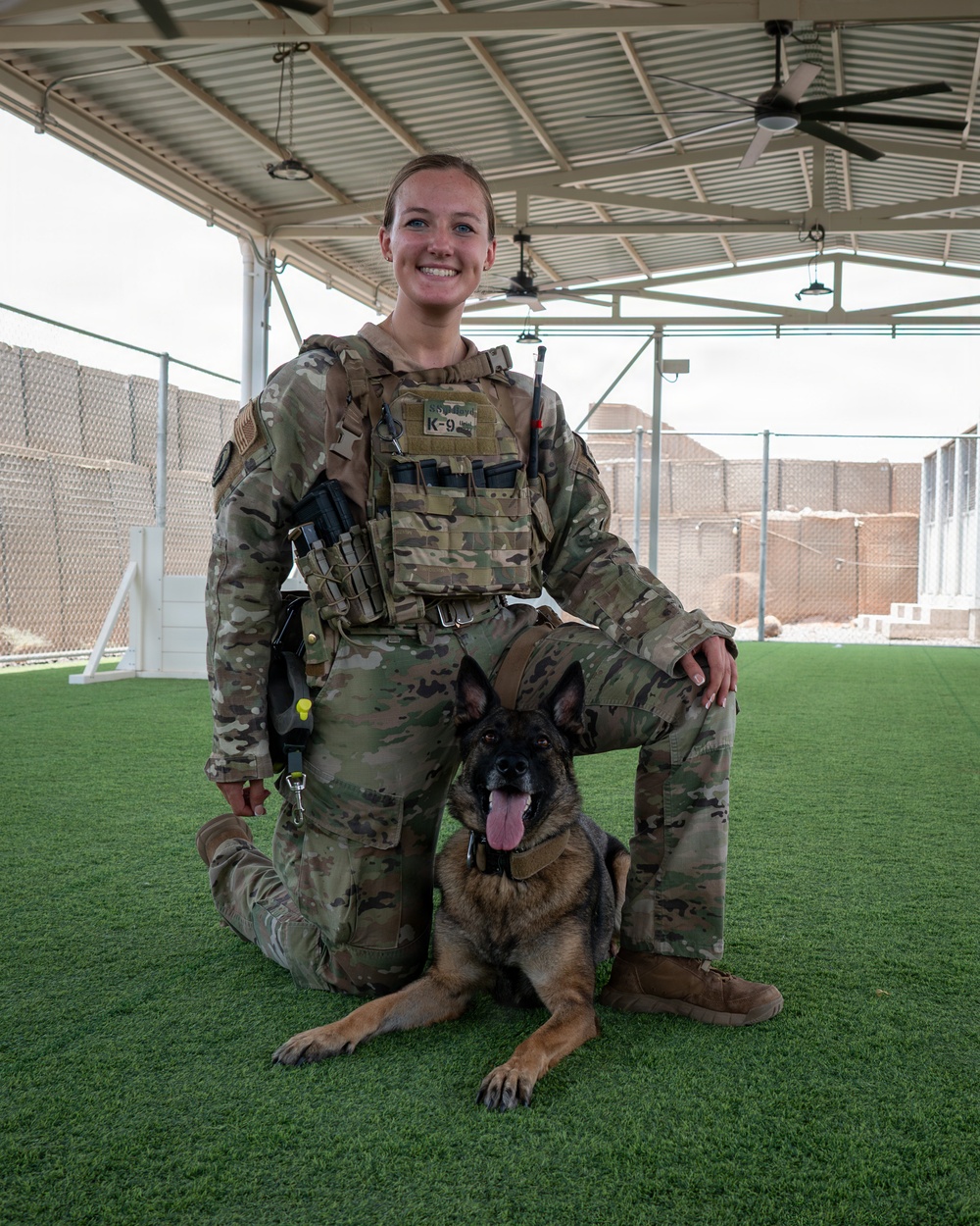 A military service member in full MultiCam/OCP camouflage uniform and tactical plate carrier vest poses with a military working dog (German Shepherd) on artificial turf under a covered outdoor area. The handler is kneeling behind the dog, which is in a down position with its tongue out. The setting appears to be a forward operating base or military installation kennel facility.